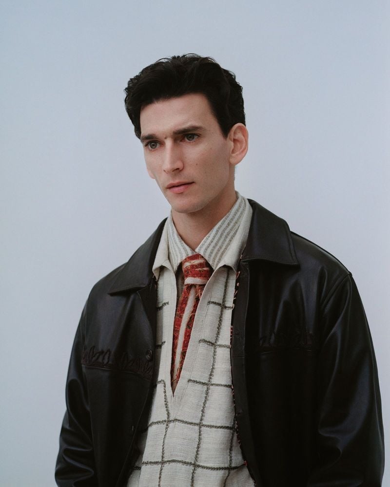 Thibaud Charon in close portrait wearing a Kartik Research black leather jacket over a cream windowpane handloom knit top and a striped shirt with a red paisley scarf at the neck, dark wavy hair, pale blue-gray studio backdrop