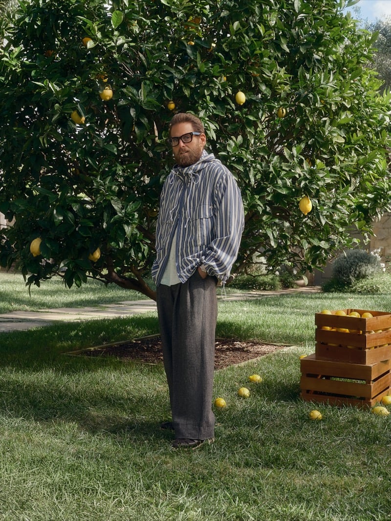 Hill stands in a blue and white striped shirt, charcoal pleated trousers, and brown loafers beside a wooden lemon crate
