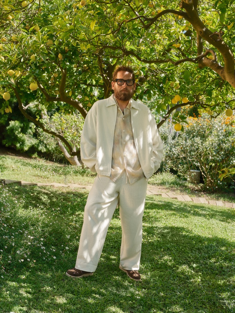 Hill stands in a cream textured bomber jacket over an ivory mesh shirt with cream wide-leg trousers and brown leather sandals on a sunlit lawn
