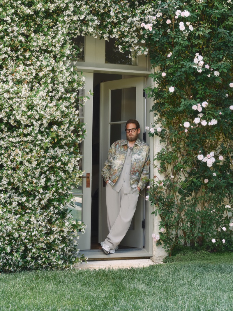 Hill leans in a doorway framed by jasmine and climbing roses, wearing a floral embroidered jacket with taupe pleated trousers