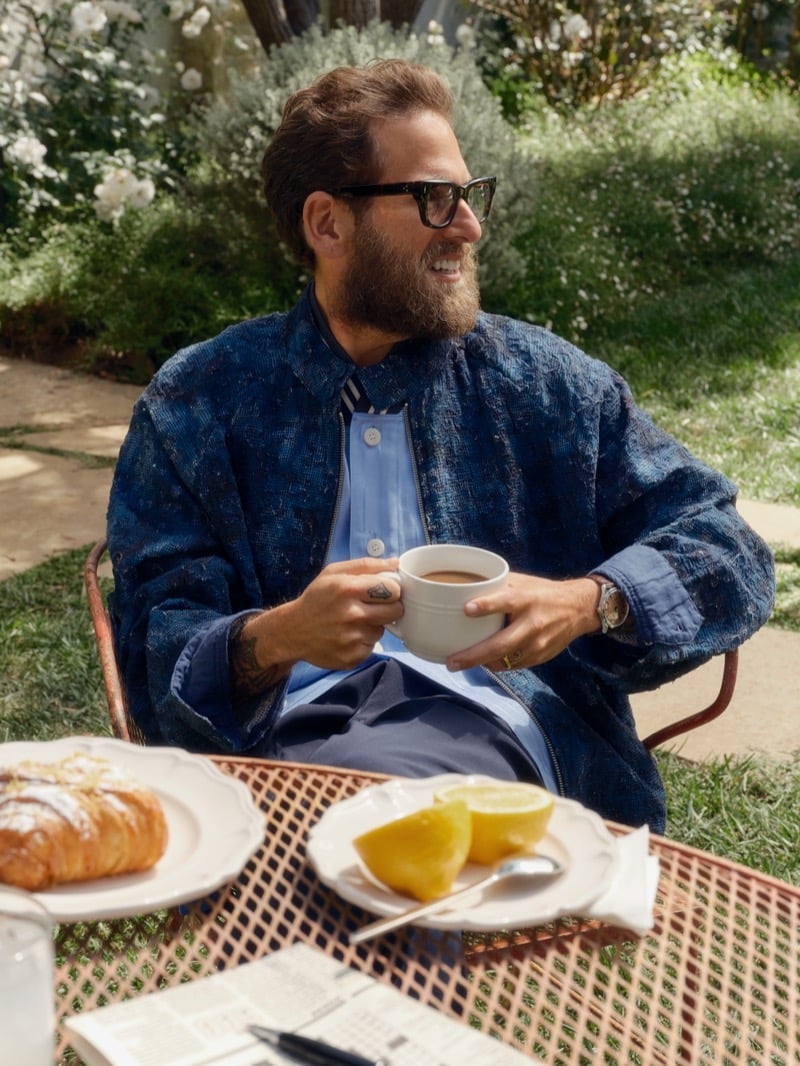Hill holds a white coffee cup at a garden breakfast table set with a croissant and sliced lemons, wearing an indigo embroidered jacket over a sky blue shirt
