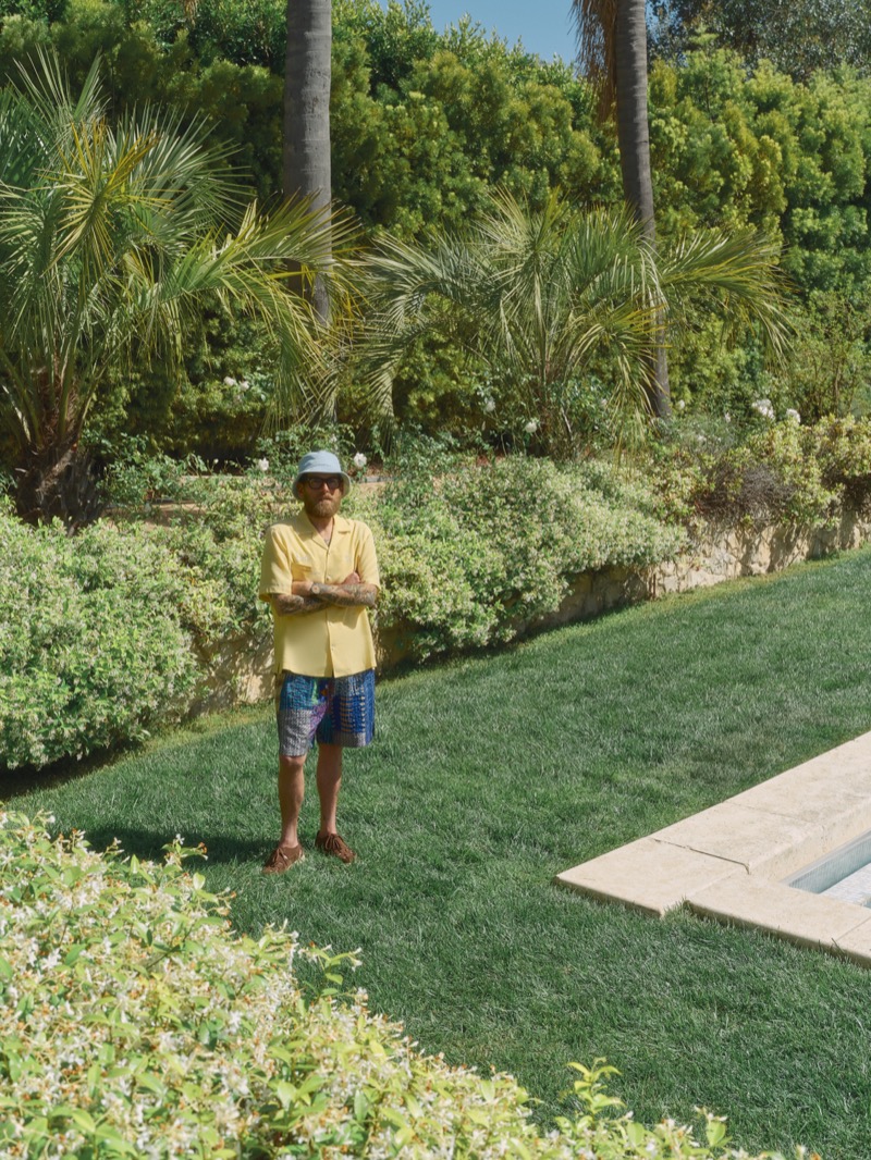 Hill stands on a lawn near a pool in a pale yellow camp shirt, printed striped shorts, a powder blue terry bucket hat, and brown sandals