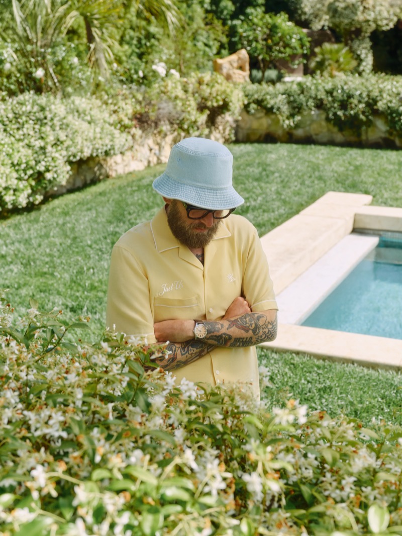 Hill crosses tattooed arms in a pale yellow camp collar shirt and a powder blue terry bucket hat beside a stone-edged pool