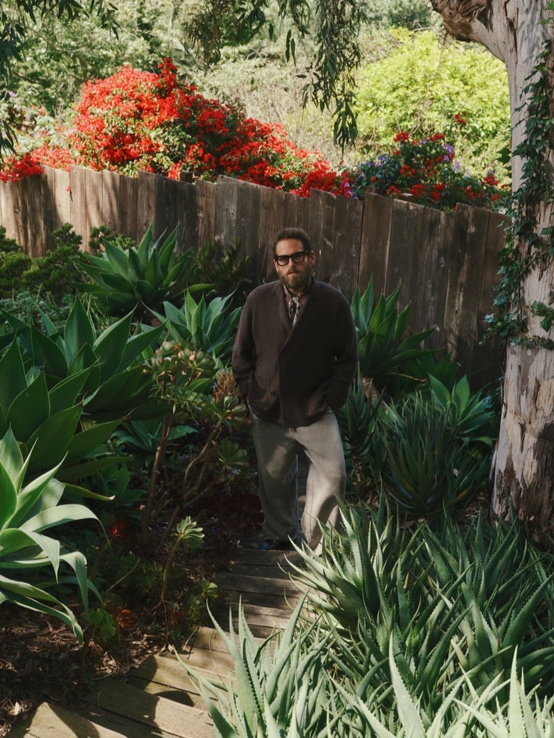 Hill walks a garden path lined with agave and red bougainvillea, wearing a dark brown jacket with oat pleated trousers and black square glasses