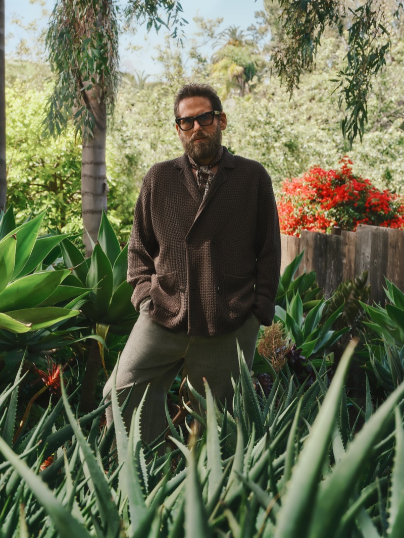 Hill stands among agave plants in a brown waffle-knit cardigan over a patterned button-up shirt, with olive pleated trousers