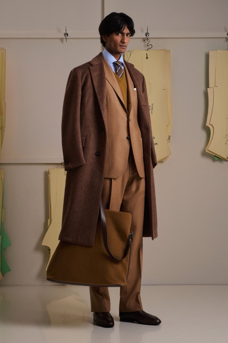 Rishi Robin wears a brown tweed single-breasted overcoat over a camel three-piece suit with an ochre sweater, blue shirt, and striped tie