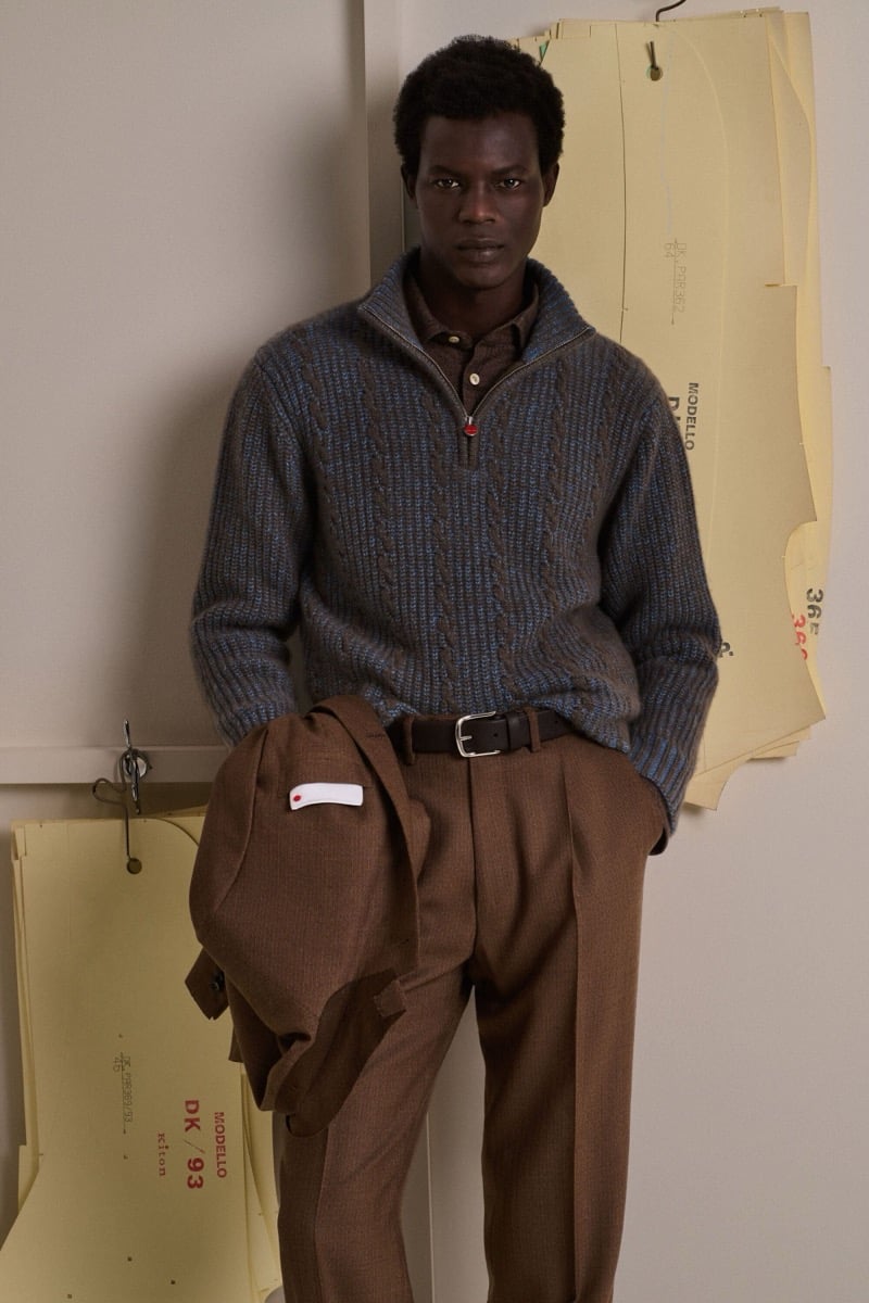 Mohammed Abubakar wears a blue and gray cable-knit half-zip sweater over a brown shirt with brown wool trousers, holding a matching brown jacket
