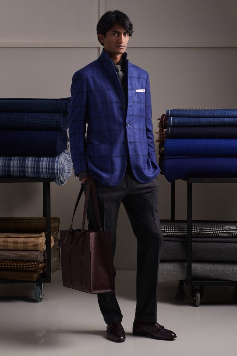 Rishi Robin wears a blue windowpane check jacket over a gray half-zip sweater with gray flannel trousers, holding a burgundy leather tote