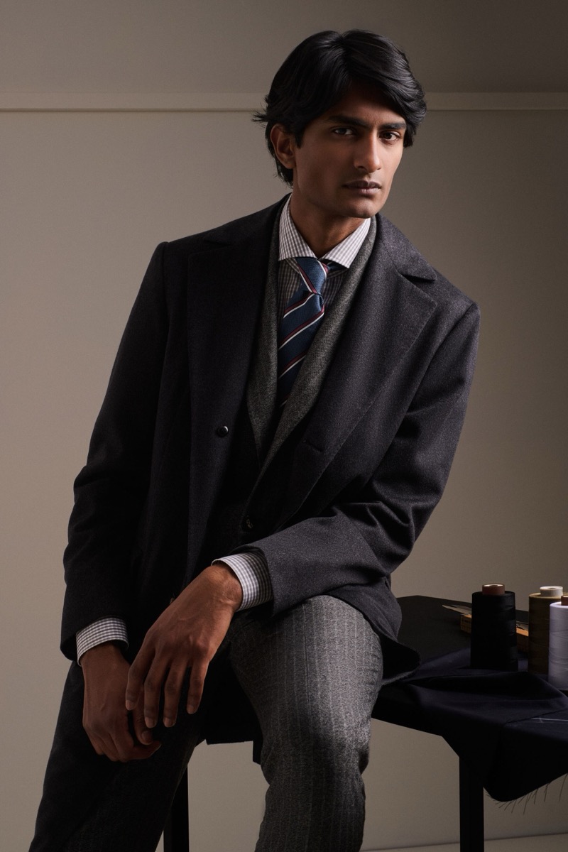 Rishi Robin wears a charcoal wool overcoat over a gray three-piece suit with a check shirt and a navy and red striped tie