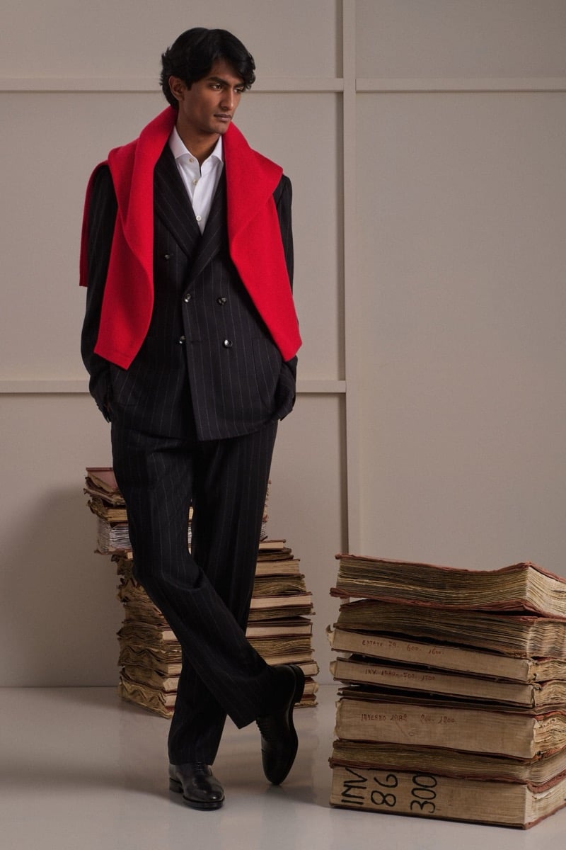 Rishi Robin wears a navy pinstripe double-breasted suit over a white shirt with a bright red sweater draped over the shoulders
