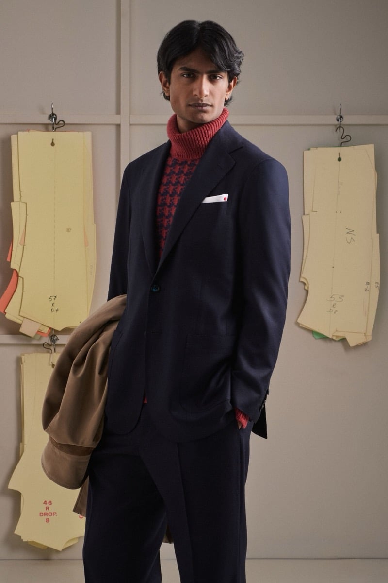 Rishi Robin wears a navy soft tailored suit over a red and navy houndstooth turtleneck, carrying a camel suede coat