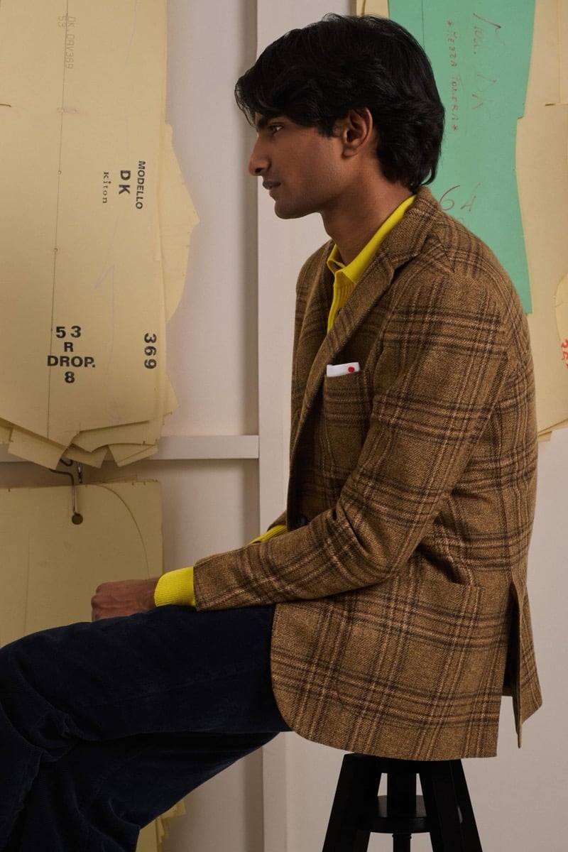 Rishi Robin sits in profile in a tan plaid wool jacket over a bright yellow polo with navy corduroy trousers