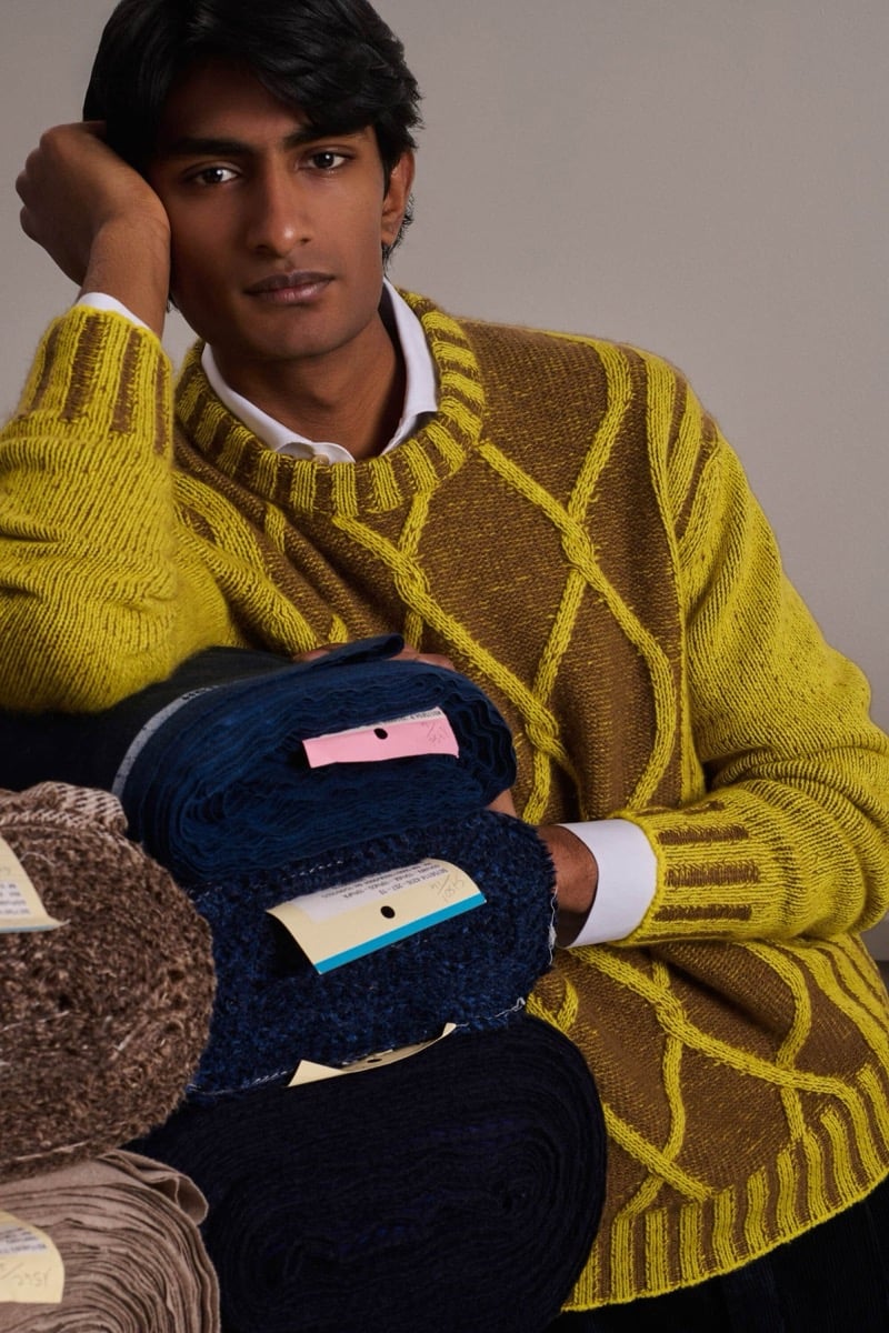 Rishi Robin wears a chartreuse and brown cable-pattern knit sweater over a white shirt, leaning against rolled cashmere bolts