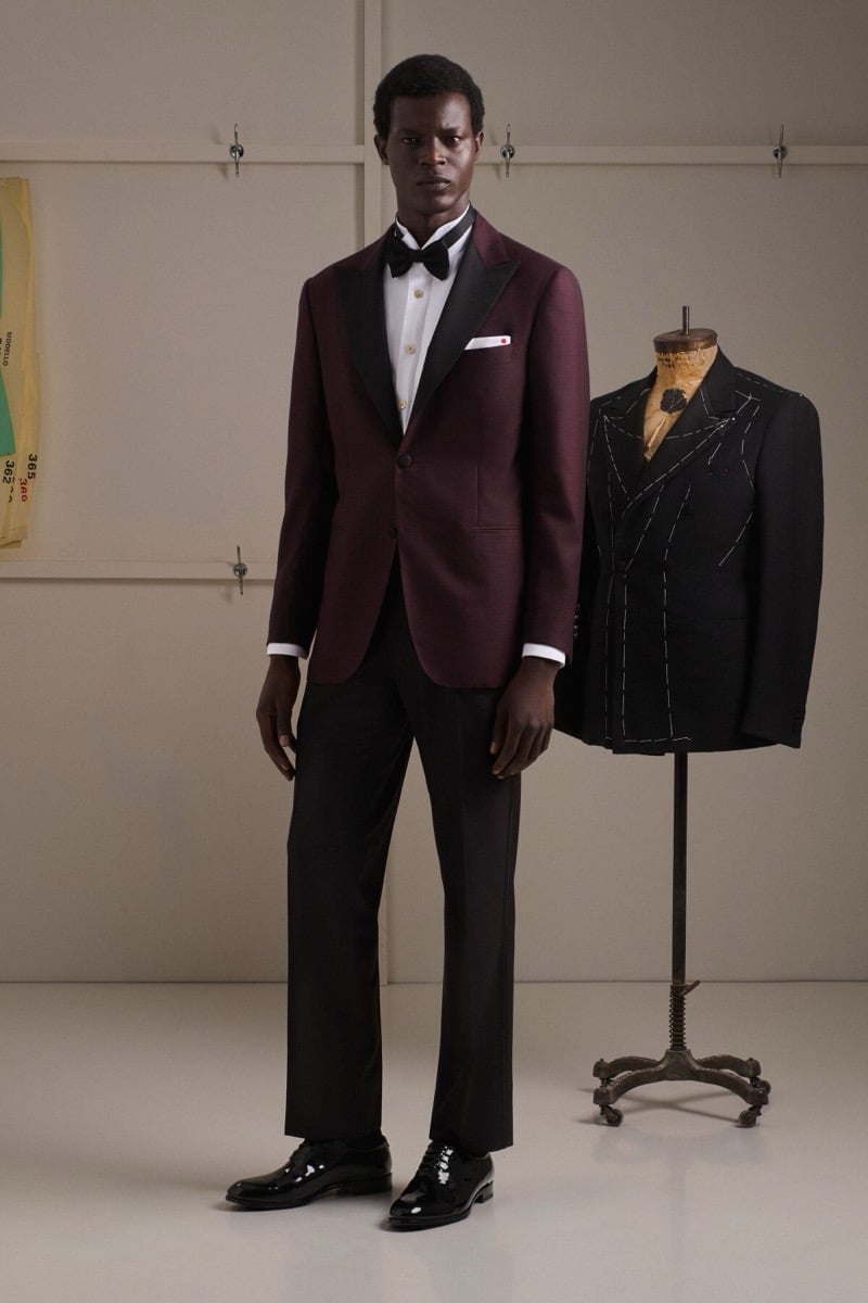 Mohammed Abubakar wears a burgundy peak-lapel dinner jacket with a black contrast lapel, a black bow tie, and black trousers, beside a half-tailored basted black dinner jacket on a mannequin