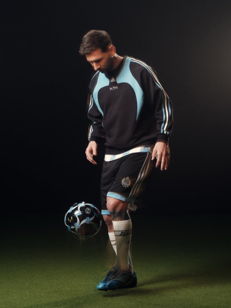Lionel juggles a chrome and black football off his right knee in a navy crewneck sweatshirt with sky blue inset panels and white adidas three-stripes, matching navy shorts with a Kith crest, knee-high tube socks and bright blue F50 cleats, on a darkened turf set