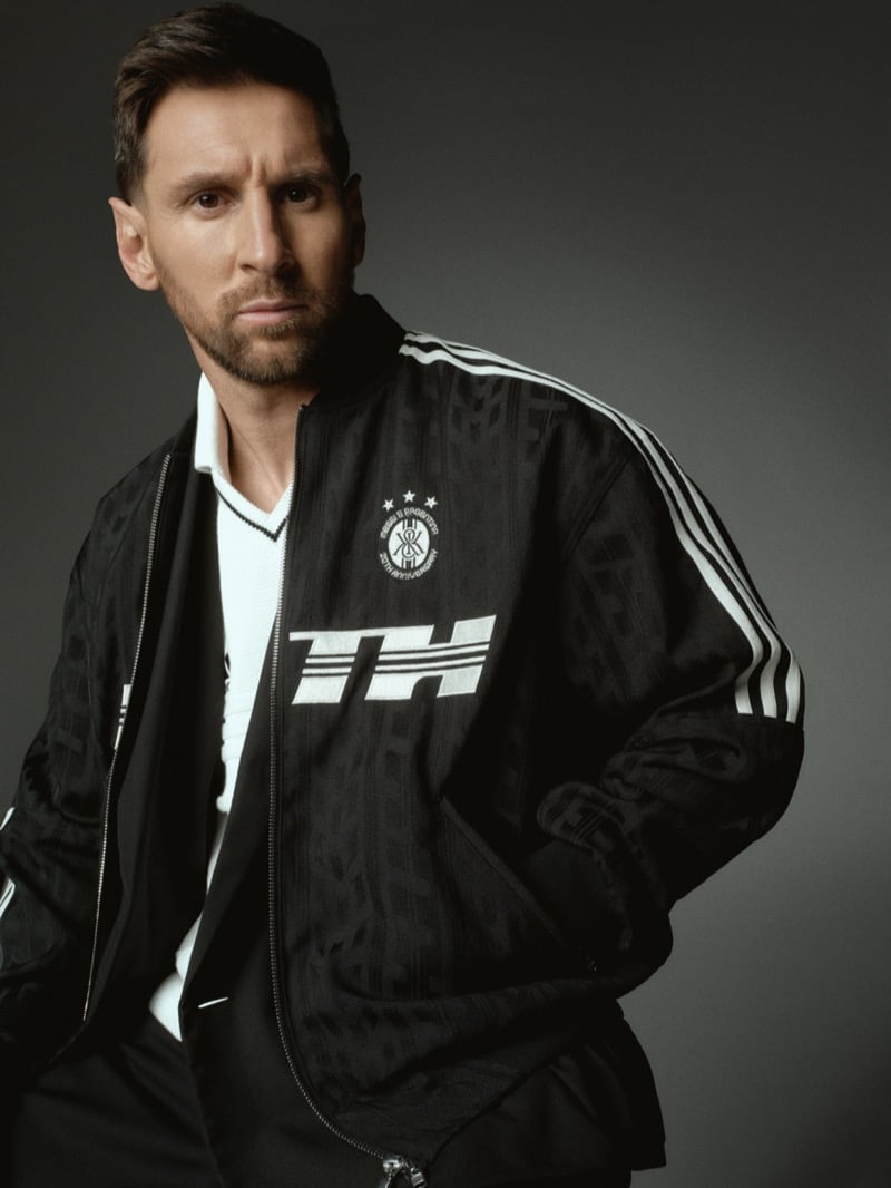 Lionel poses against a slate backdrop in a black tonal jacquard track jacket with white adidas three-stripes down the arms, a white block-letter chest graphic and a circular Kith x adidas anniversary crest at the heart, layered over a white knit polo and black trousers