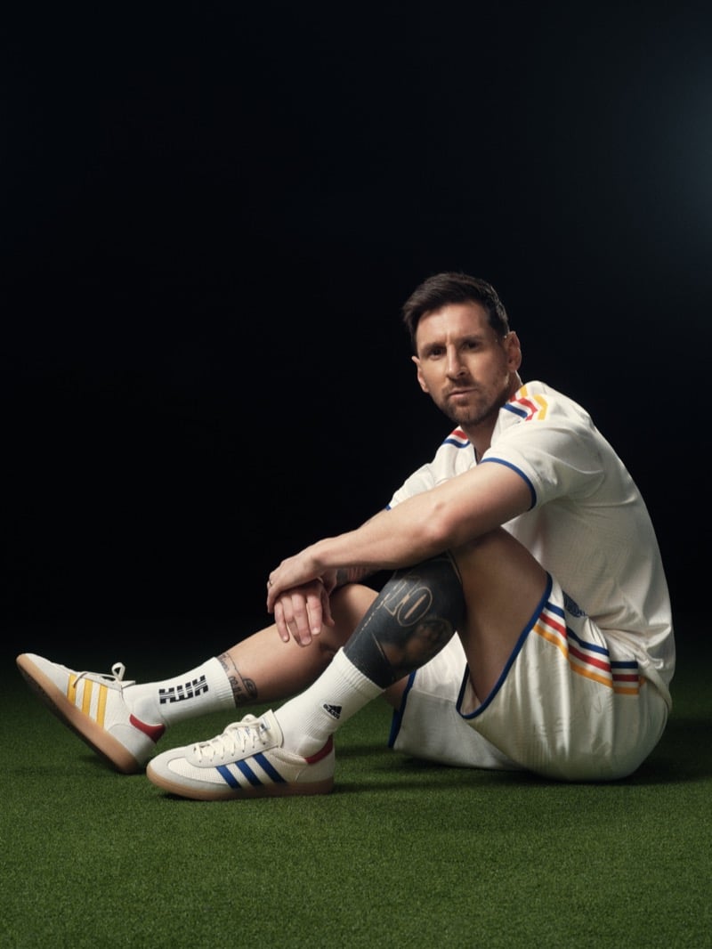 Lionel sits cross-legged on turf in a cream training jersey and matching shorts trimmed in red, blue, and yellow at the collar, sleeves, and hems, white KITH-lettered tube socks, and a pair of off-white adidas Samba sneakers with three-stripe inserts in red, blue, and yellow on each foot