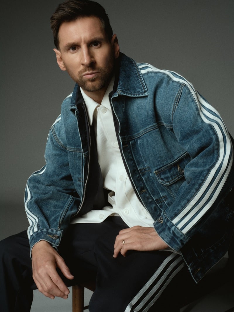 Lionel sits on a stool in an oversized washed indigo adidas denim trucker jacket with white three-stripes down both arms and a small Kith heart patch, layered over a cream knit overshirt and dark trousers with white side stripes, hands resting on his knees