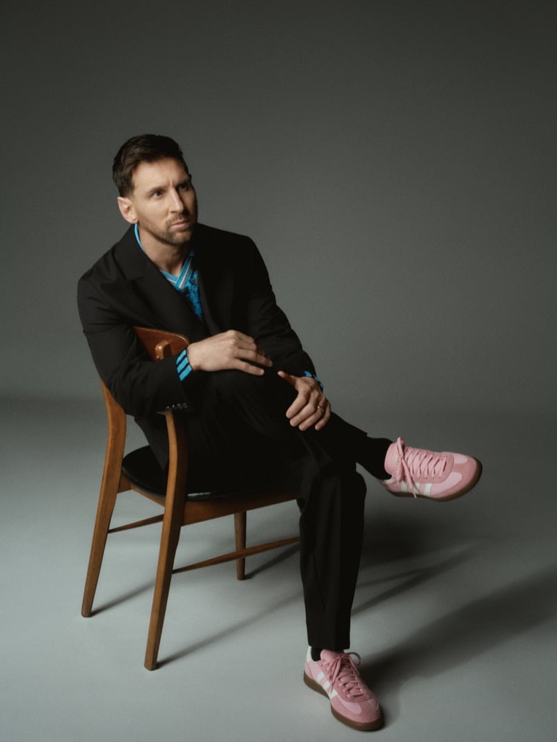 Lionel leans back in a bentwood chair in a relaxed black single-breasted blazer and matching trousers worn over an Argentina-striped knit, crossed at the ankle to show baby pink suede adidas Gazelle sneakers