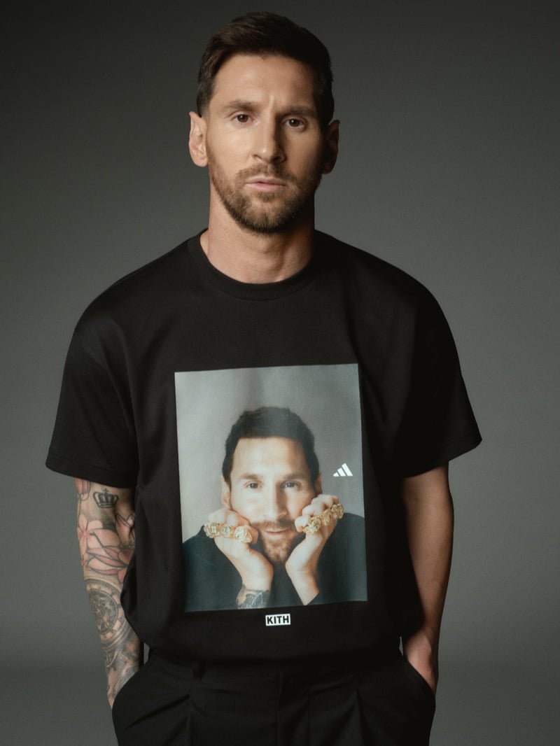 Lionel faces forward in a black cotton t-shirt printed with a faded portrait of himself resting his ringed knuckles against his chin, a small white adidas trefoil at the upper right of the print and a Kith woven tab at the lower edge, paired with black pleated trousers and a sleeve of arm tattoos visible at left