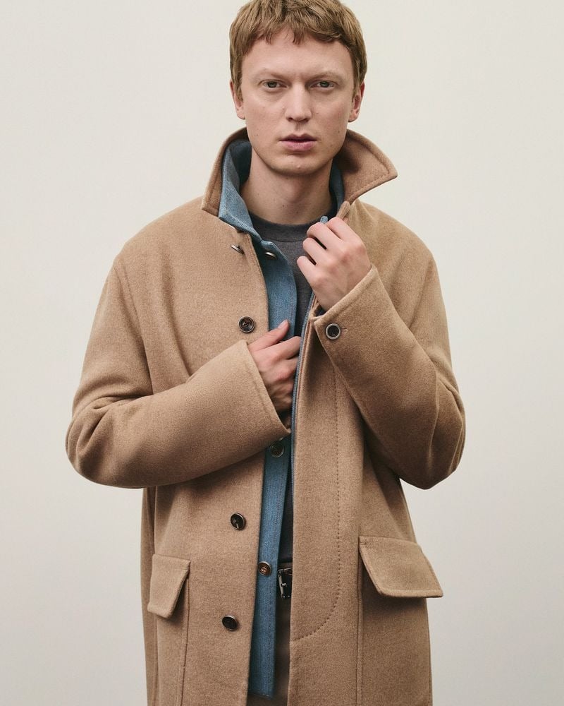 Jonas stands against a pale studio backdrop in a camel cashmere stand-collar coat layered over a blue denim jacket and a grey crewneck