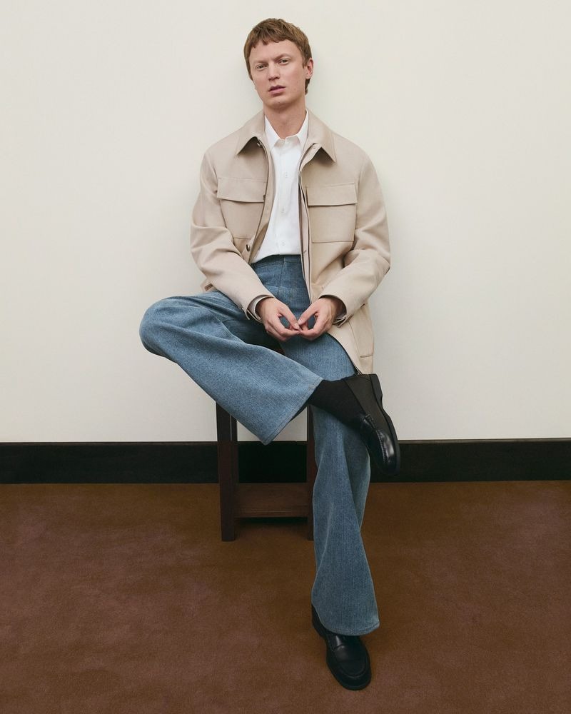 Jonas sits on a dark wood stool against a cream wall in a boxy cream utility jacket with chest pockets, a white shirt underneath, faded blue wide-leg denim, and black loafers