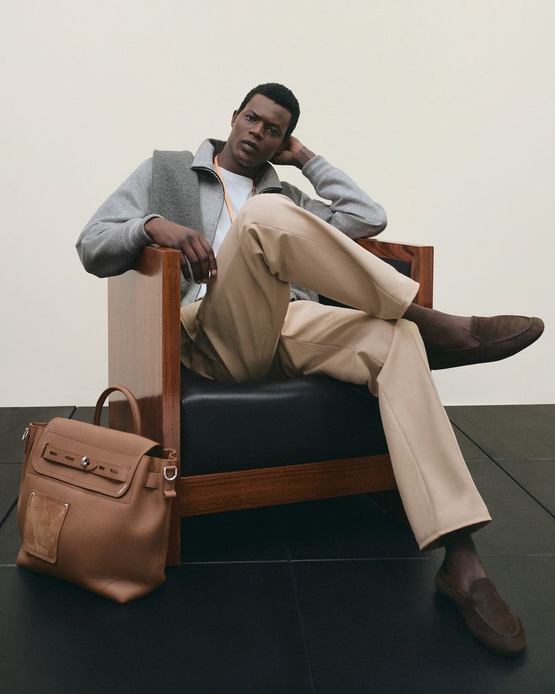 Mohammed reclines in a mid-century brown wood chair with a black leather cushion, wearing a gray wool zip bomber over a white tee, beige tailored trousers, and brown suede loafers, a tan leather Louis Vuitton Steamer 30 bag set on the floor beside him