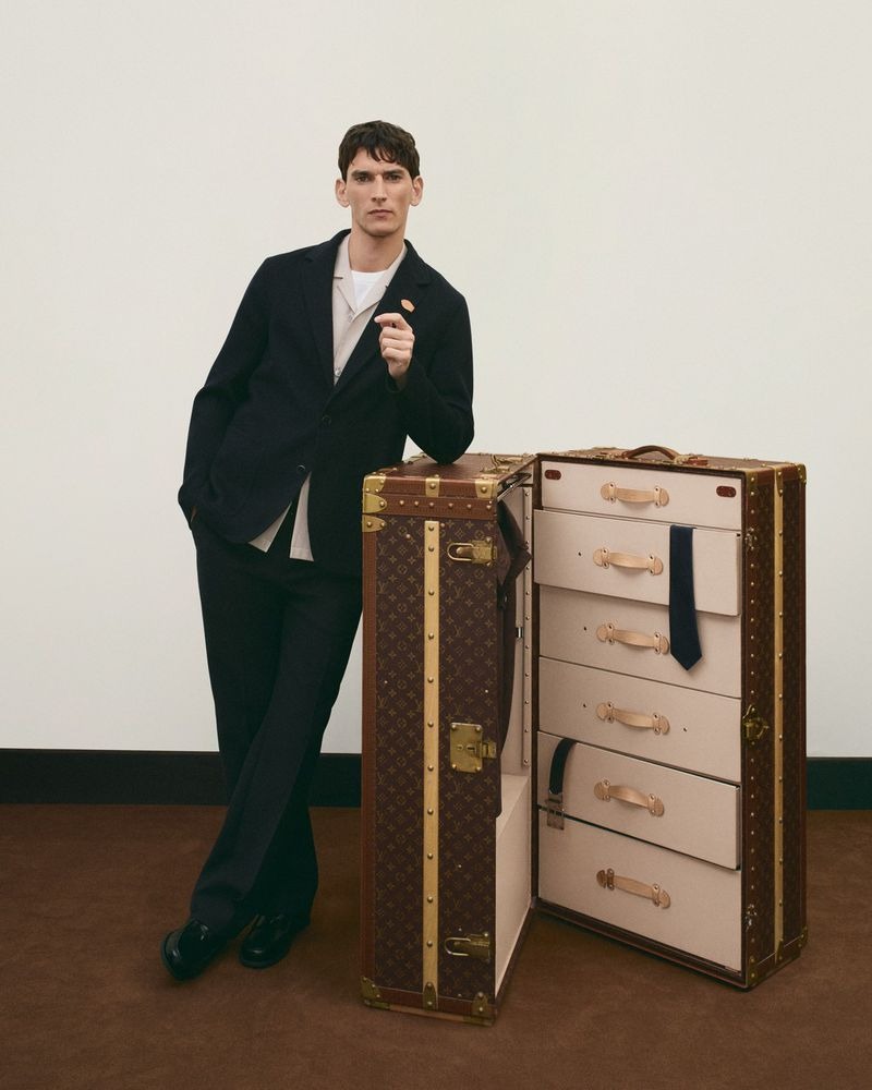 Thibaud stands beside an open Louis Vuitton monogram wardrobe trunk revealing ivory canvas drawers with tan leather handles and a navy tie hung inside, wearing a blazer open over a beige shirt, white tee, dark wide trousers, and black loafers