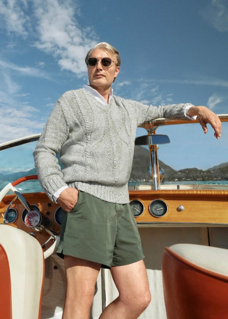Mads Mikkelsen stands at the wheel of a varnished Riva Aquarama wooden speedboat on Lake Maggiore, wearing sunglasses, a marled gray cable-knit V-neck sweater over an open white shirt, and olive green tailored swim shorts, the lake and mountains stretching behind him
