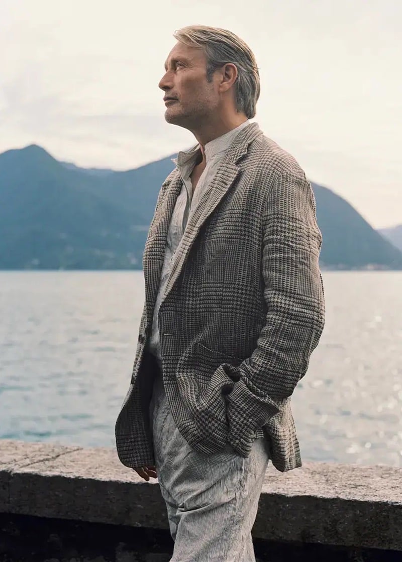 Mads Mikkelsen stands in profile on a stone lakeside promenade at Lake Maggiore, wearing a glen check Oasi Lino linen blazer in soft brown and ivory tones over a cream linen shirt with pinstripe trousers, mountains rising across the water behind him