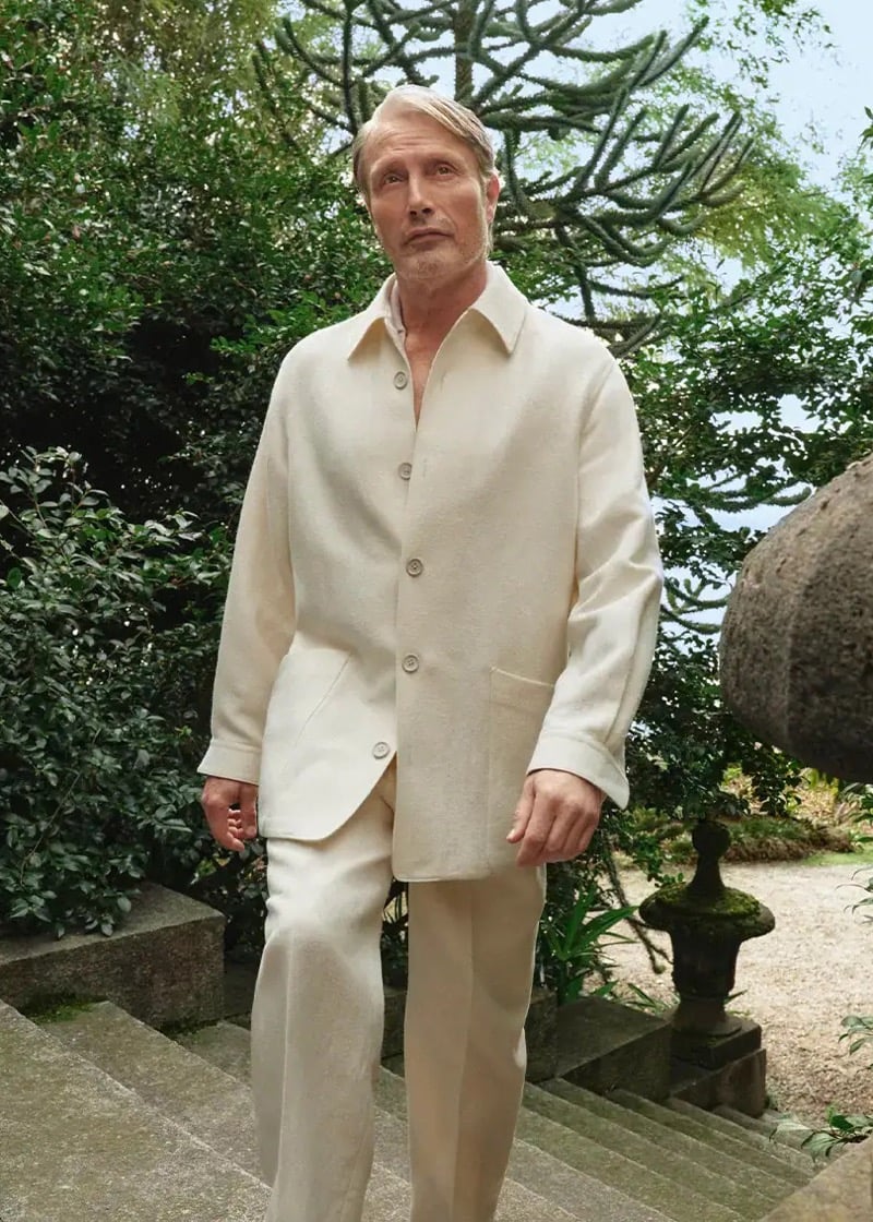 Mads Mikkelsen walks down stone garden steps surrounded by Mediterranean foliage at Lake Maggiore, wearing an off-white textured shirt jacket buttoned over matching trousers in a soft natural fabric