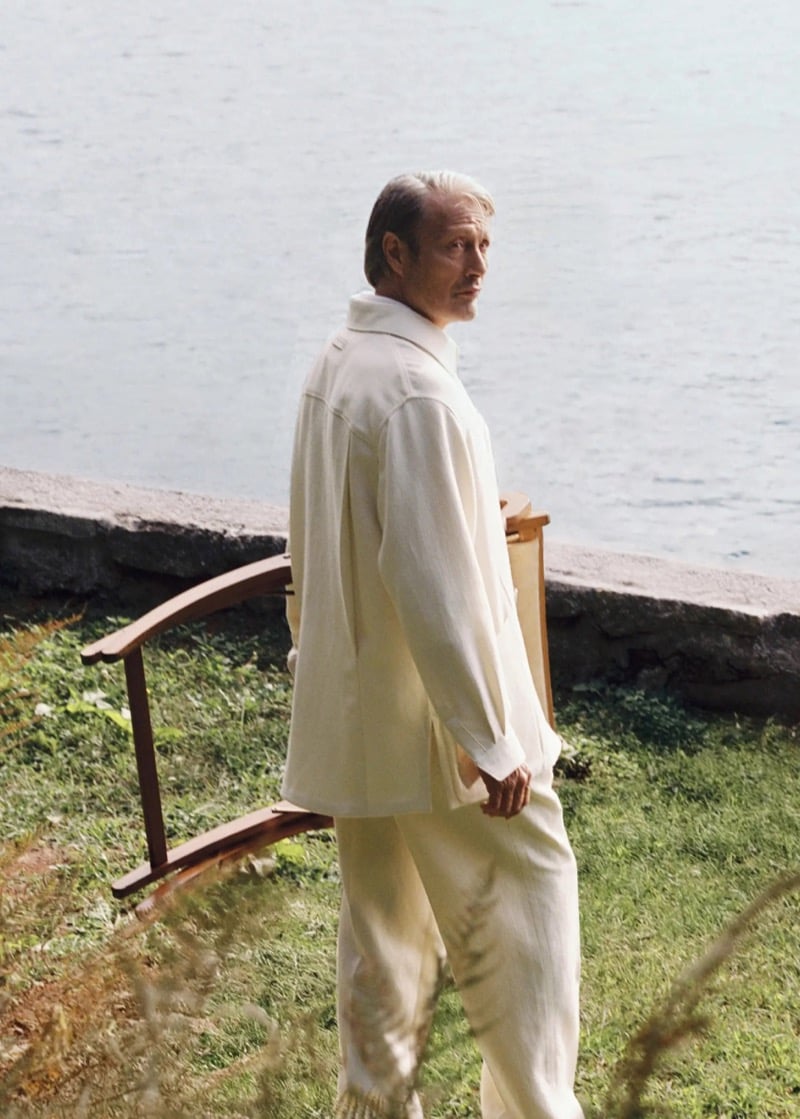 Mads Mikkelsen pauses on a grassy lawn beside Lake Maggiore, glancing over his shoulder while wearing a cream linen overshirt with matching tailored trousers, a wooden footbridge and the still water visible behind him