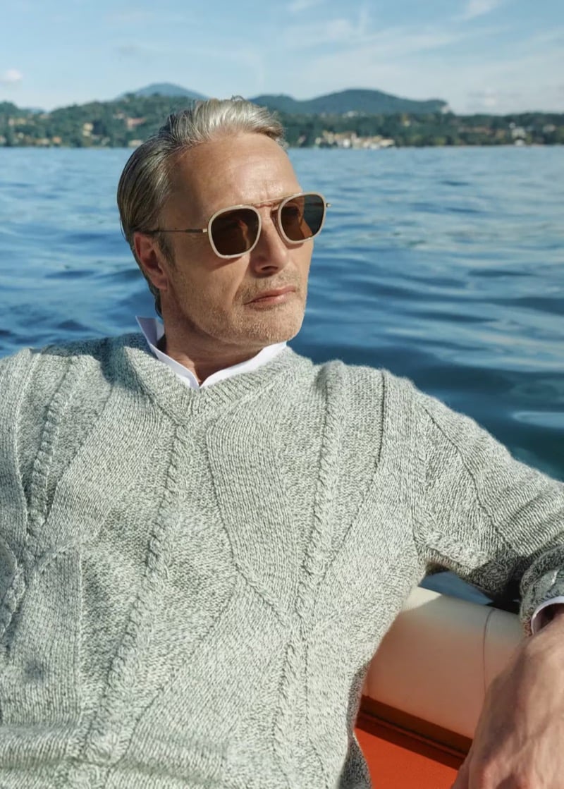 Mads Mikkelsen photographed in close portrait aboard a speedboat on Lake Maggiore, wearing pale sunglasses, a marled gray cable-knit V-neck sweater, and an open white linen shirt, the lake stretching out behind him