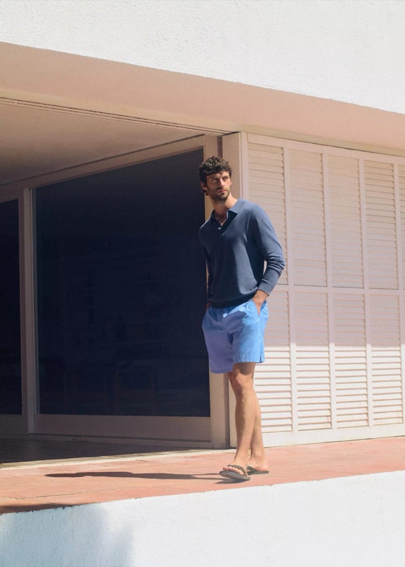 Matthew stands in flip-flops in the sunlit doorway of a Mediterranean villa wearing a navy fine-gauge knit polo and cornflower blue drawstring swim shorts