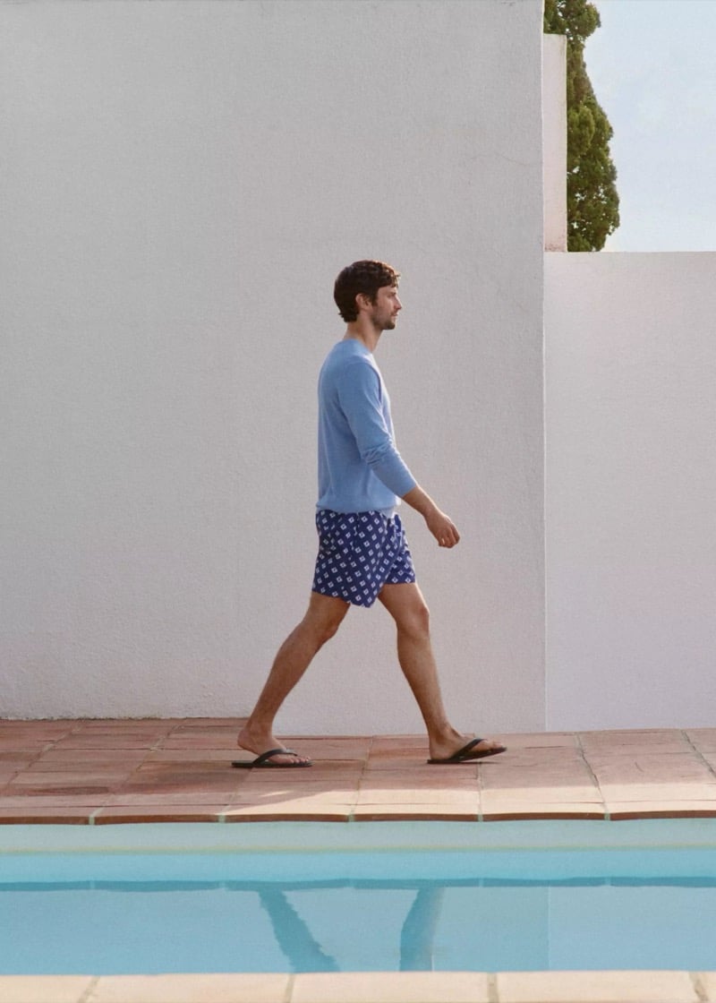 Matthew walks past a tiled pool in profile wearing a powder blue crew-neck cotton sweater and navy and white tile-print swim shorts with flip-flops