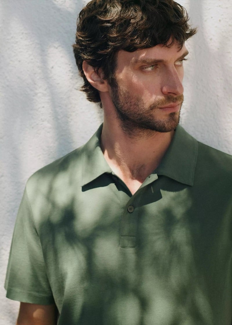 Close-up of Matthew with curly hair and a beard in a sage green cotton polo shirt against a dappled white wall
