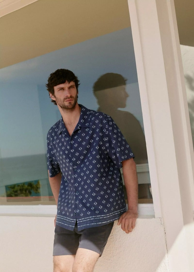Matthew leans against a villa window reflecting the Mediterranean in a navy bandana-print short-sleeve camp collar shirt and grey shorts