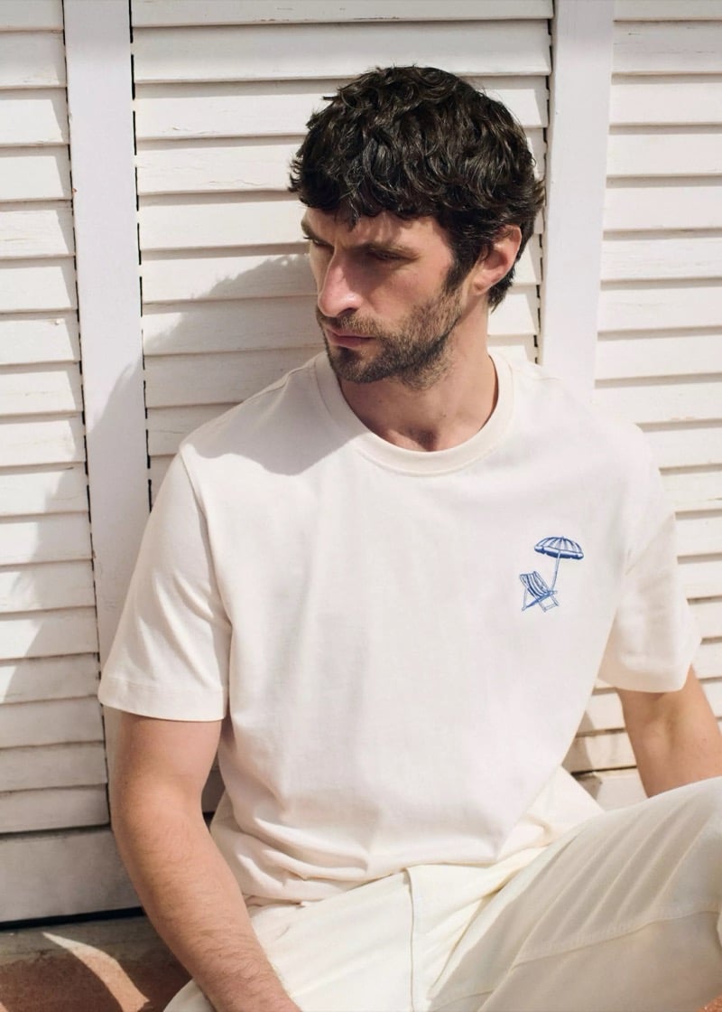 Close-up of Matthew seated against a white louvered shutter in a cream cotton t-shirt with a small blue embroidered beach chair and umbrella on the chest