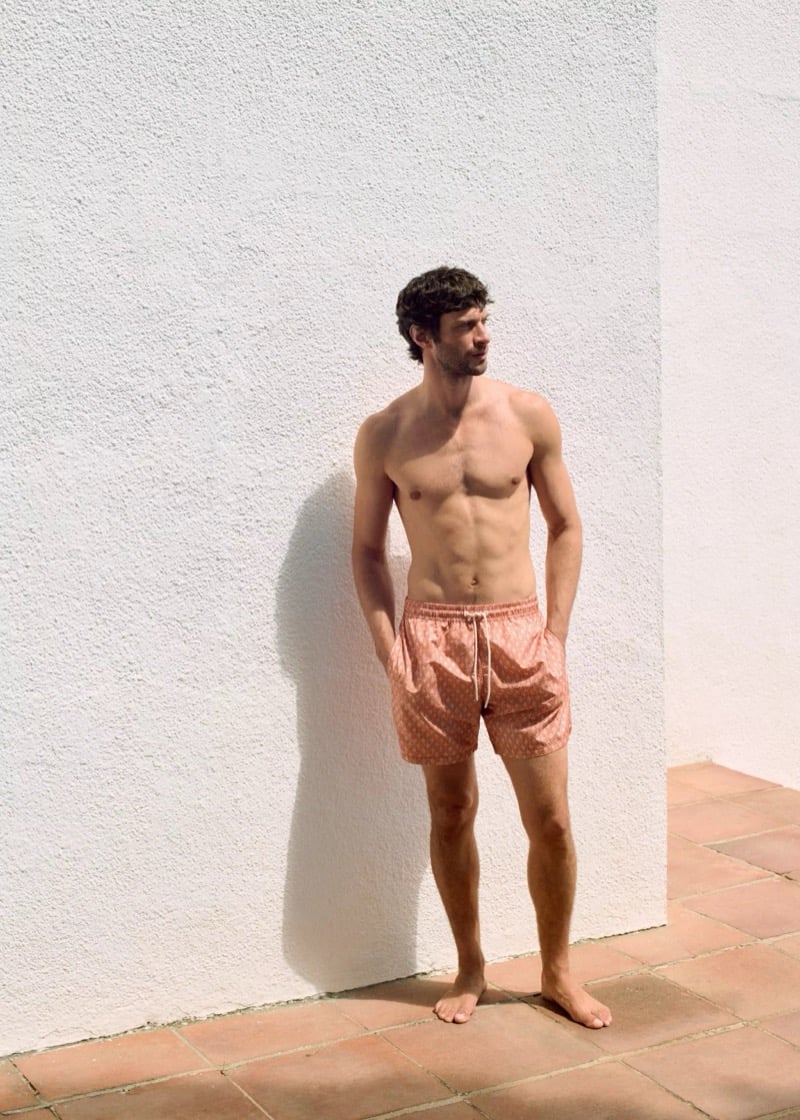 Matthew stands shirtless on terracotta tile against a white stucco wall in coral pink small-print drawstring swim shorts