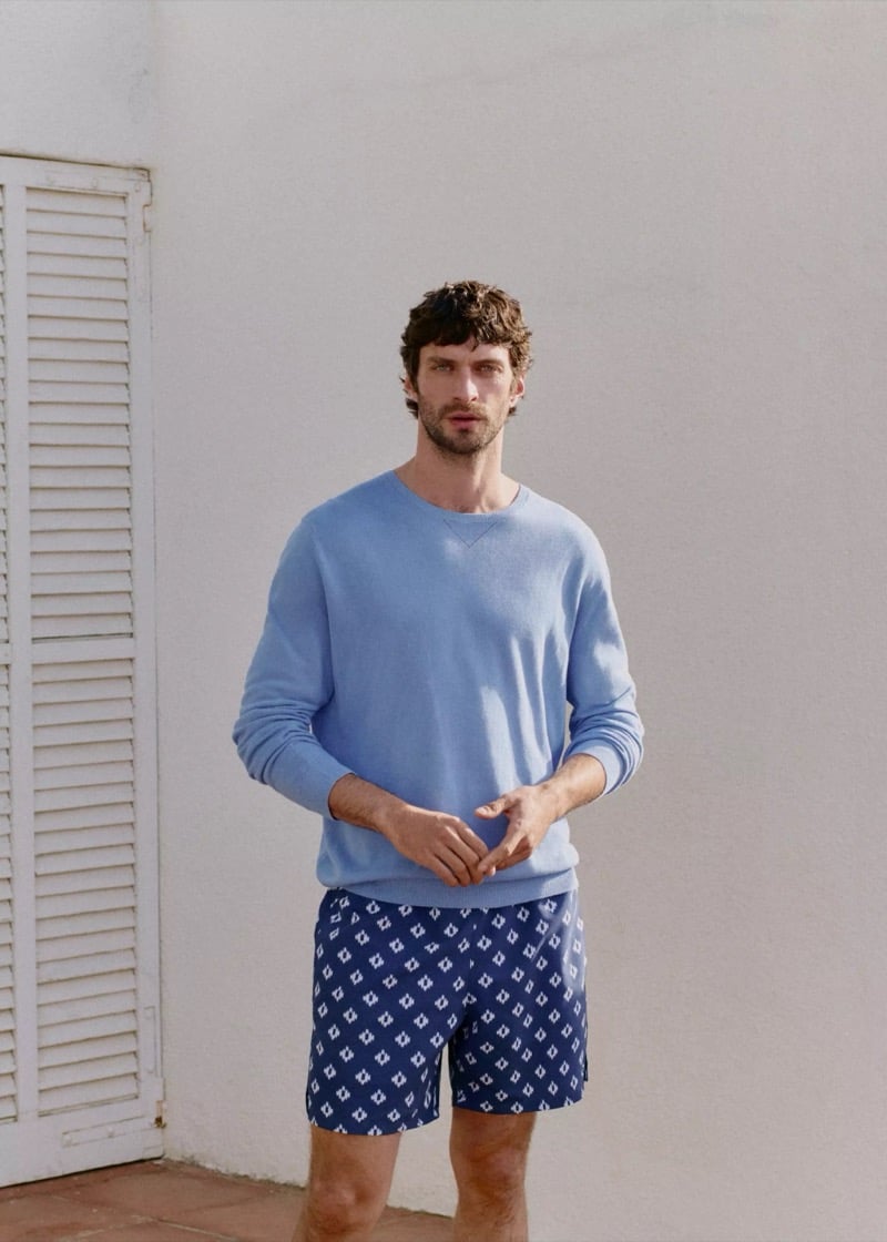 Matthew stands against a white wall and louvered shutter in a powder blue cotton crew-neck sweater and navy geometric-print swim shorts