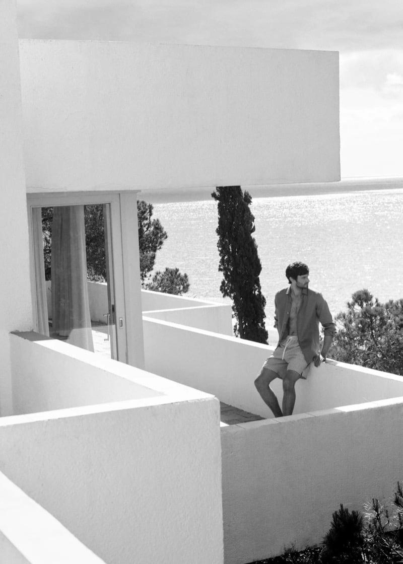 Black and white image of Matthew on a whitewashed villa terrace overlooking the Mediterranean in an open terracotta linen shirt and seersucker striped shorts