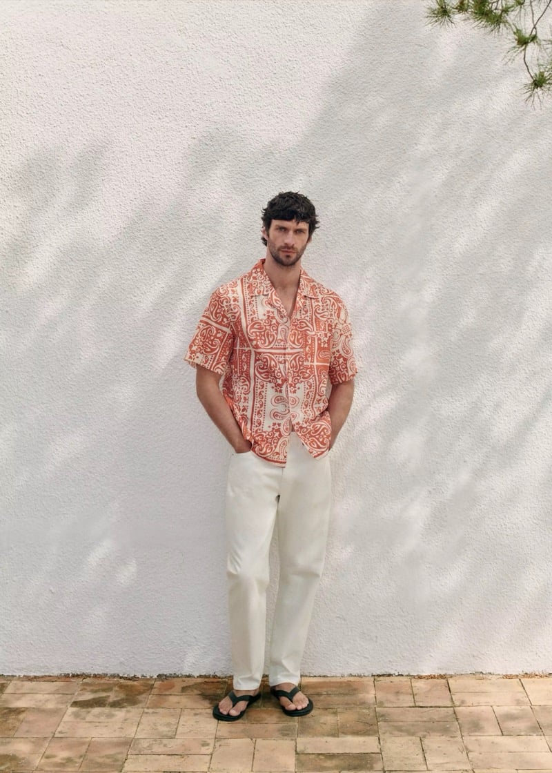 Matthew stands against a dappled white stucco wall in a coral paisley short-sleeve camp collar shirt and cream white linen trousers with slide sandals