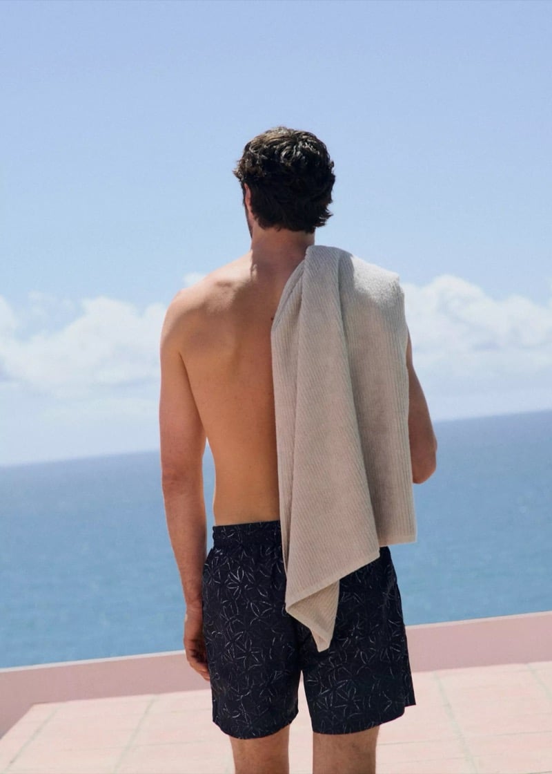 Back view of Matthew at a pool with a grey towel slung over his shoulder, the Mediterranean beyond, wearing navy printed swim shorts with a small scattered motif