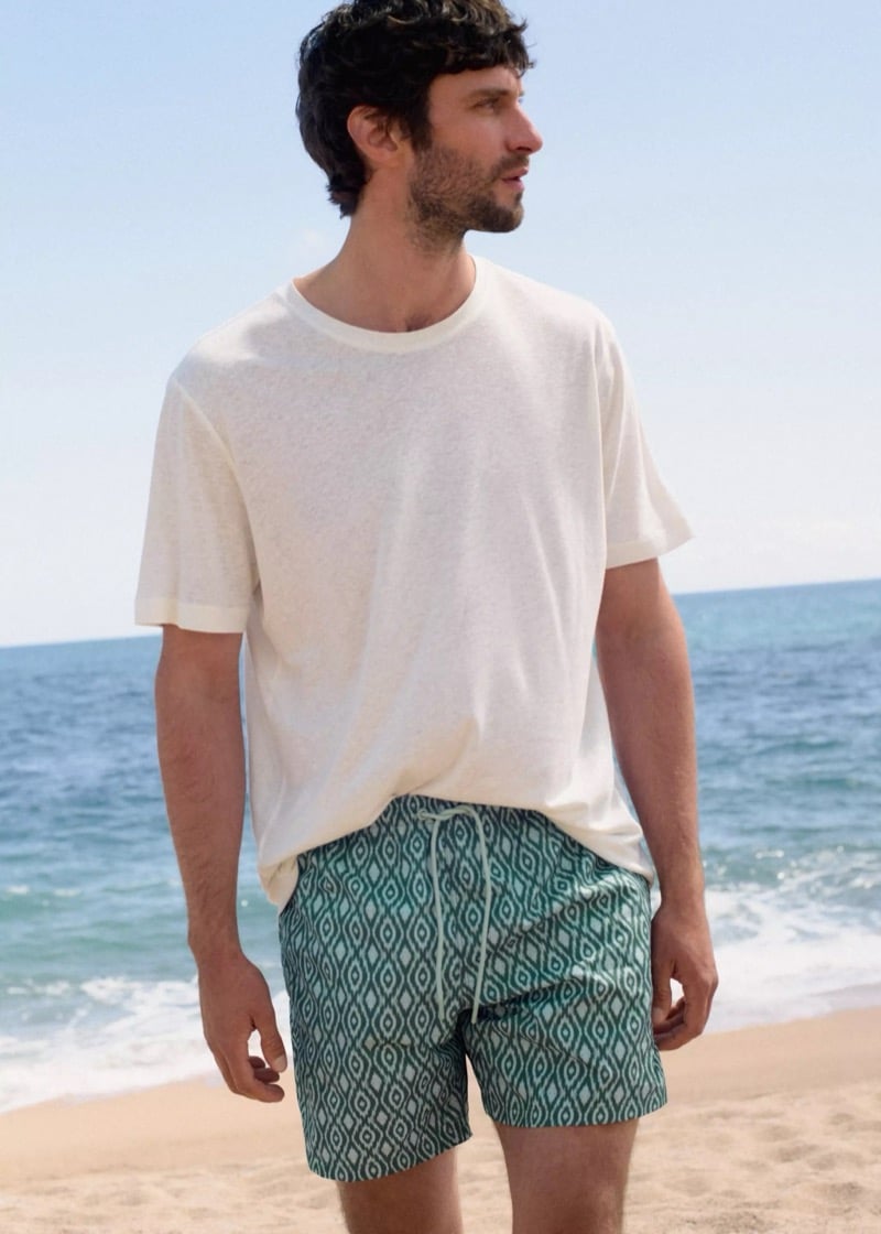 Matthew stands on the sand in front of the sea in a cream linen t-shirt and green and white ikat-print drawstring swim shorts