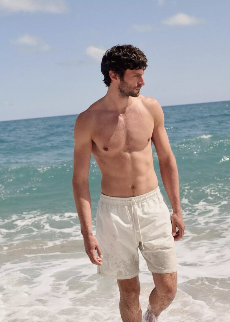 Matthew stands shirtless in the surf with the Mediterranean around him in cream seersucker drawstring swim trunks