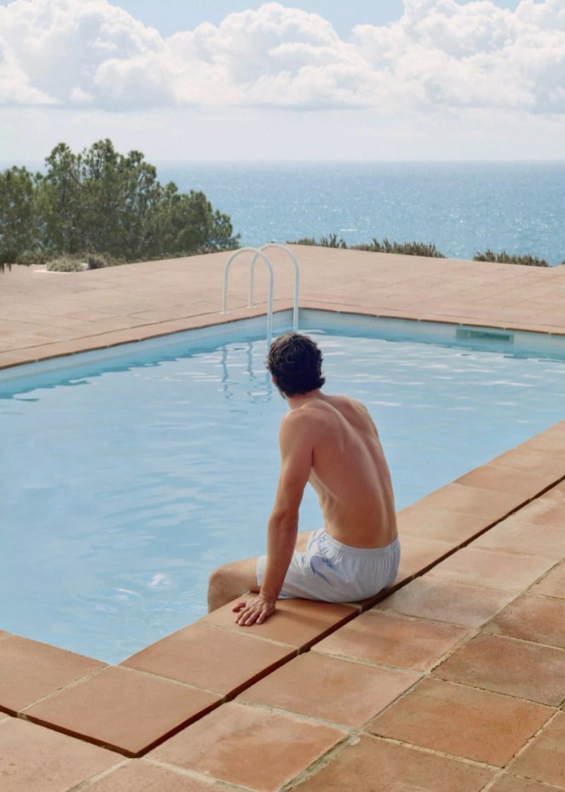 Matthew sits at the edge of a tiled pool with his back to the camera in white seersucker swim trunks, the Mediterranean beyond