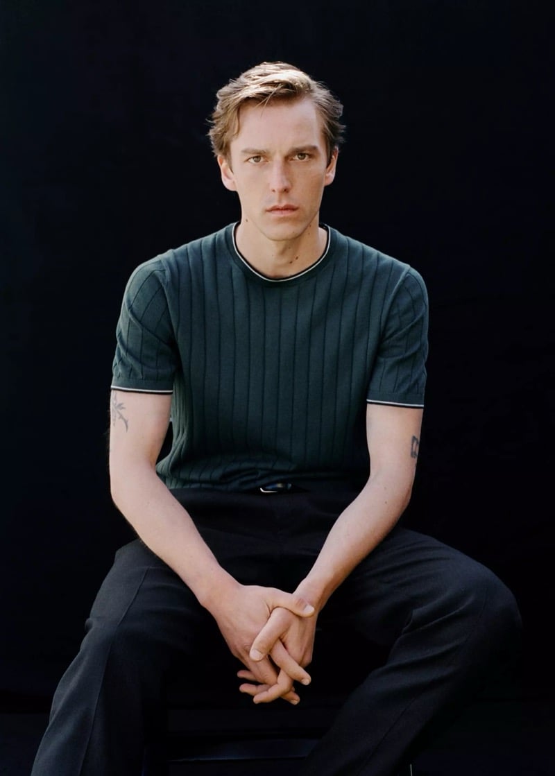 Quentin sits in a forest green ribbed crewneck knit tee with black trousers against a black backdrop