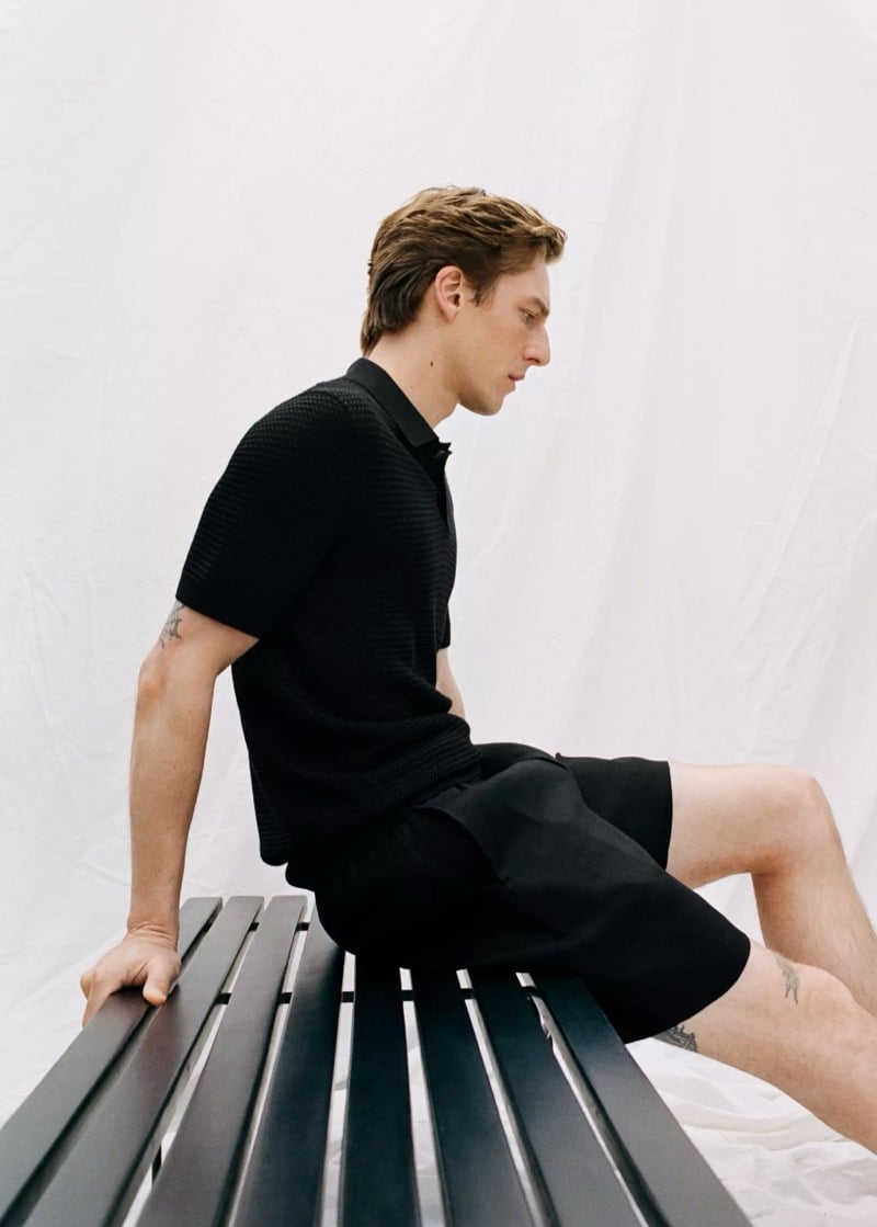 Quentin sits on a black bench in a Mango textured black knit polo and black drawstring Bermuda shorts