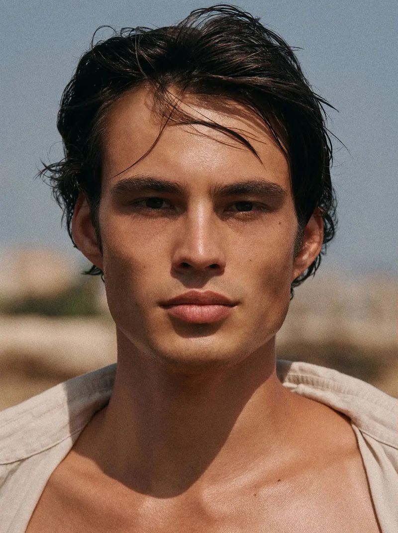 Close-up portrait of Louis with wet swept-back dark hair and a sand-colored linen shirt opening at the collarbone against a soft coastal background
