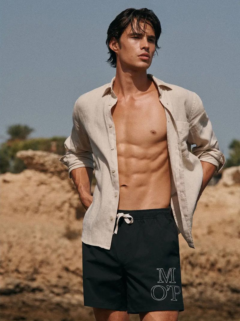 Louis wears an open sand-colored linen shirt with rolled sleeves over black swim shorts with a white M O’P monogram